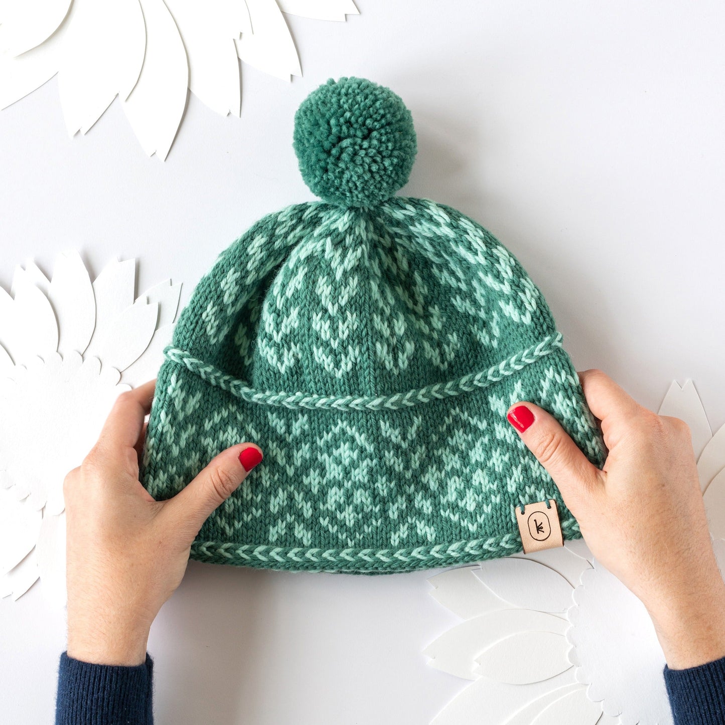 White hands with pink nail polish holding the August Hat pattern by Sarah Solomon, knit out of Germantown in 338 jade and 319 myrtle on a paper background with sunflower paper cut out shapes