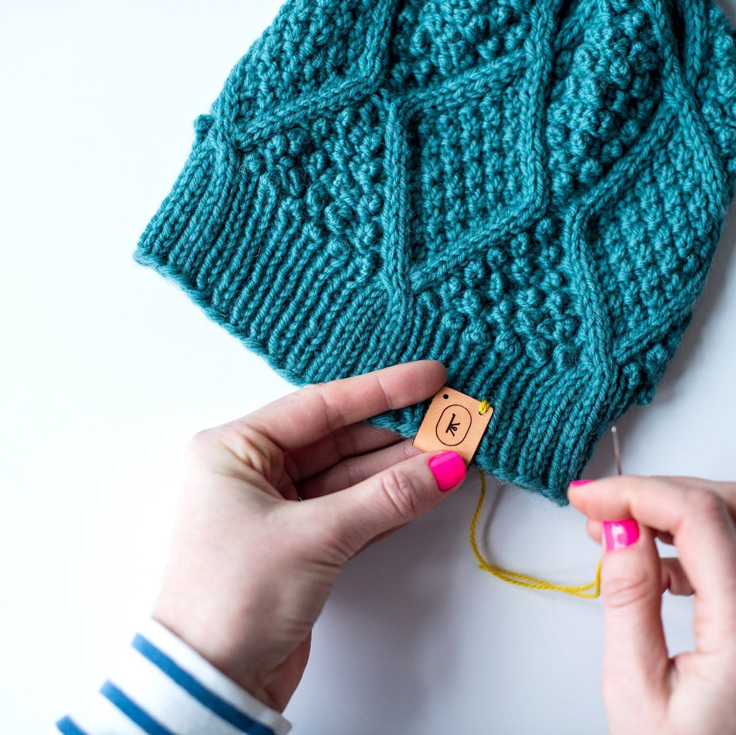 White hands with a blue and white long sleeve shirt and pink painted nails sewing a leather hat tag on the heavily bobbled and cabled January Hat, knit in Germantown in sage.
