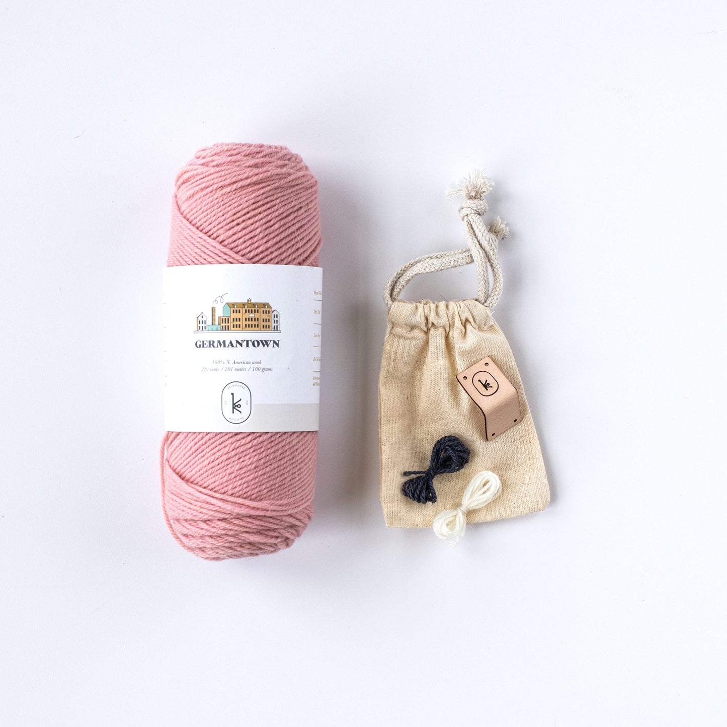 Flat lay of 687 baby pink Kelbourne Woolens Germantown yarn skein with 2 small colorful yarn butterflies and a leather tag on a small muslin bag.