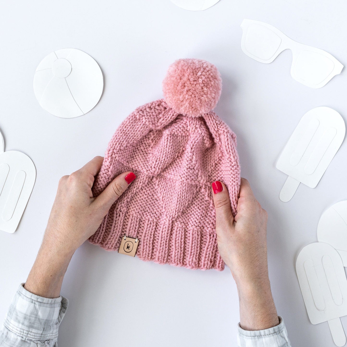 White hands with pink nail polish holding the July Hat pattern by Courtney Kelley, knit out of Germantown in 687 baby pink on a paper background with paper popsicles, sunglasses, and ball cut out shapes