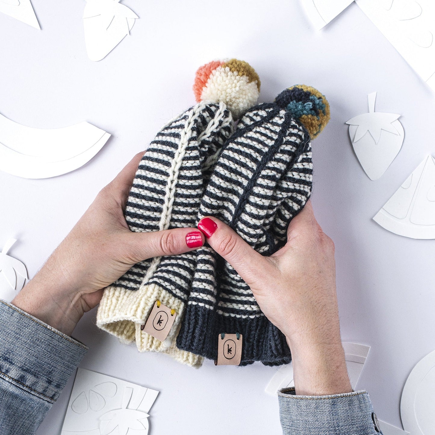 White hands with pink nail polish holding the June Hat pattern by Meghan Kelly, knit out of Germantown in 005 and black and 105 natural with colorful pompoms on a paper background with fruit paper cut out shapes