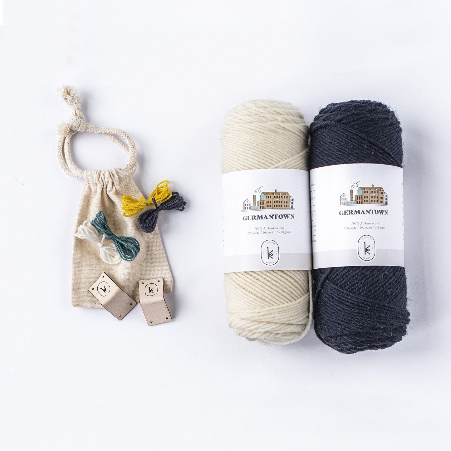 Flat lay of 105 natural and 005 black Kelbourne Woolens Germantown yarn skeins with 4 small colorful yarn butterflies and two leather tags on a small muslin bag.