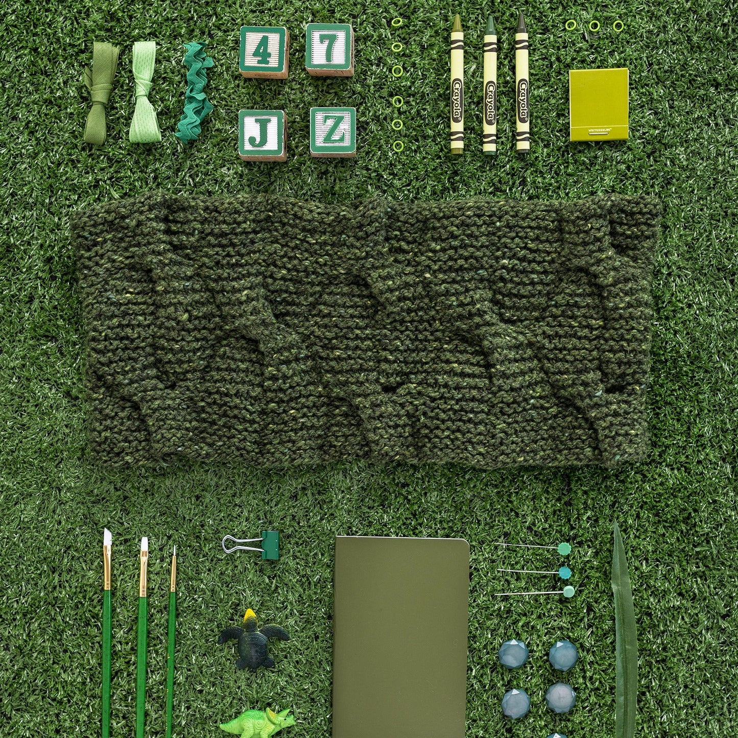 The Shamrock Cowl by Courtney Kelley knit in Kelbourne Woolens Lucky Tweed in pine green pictured on an astroturn ground surrounded by blocks, crayons, buttons, pinks, pencils, and paintbrushes neatly arranged in a "knolling" fashion