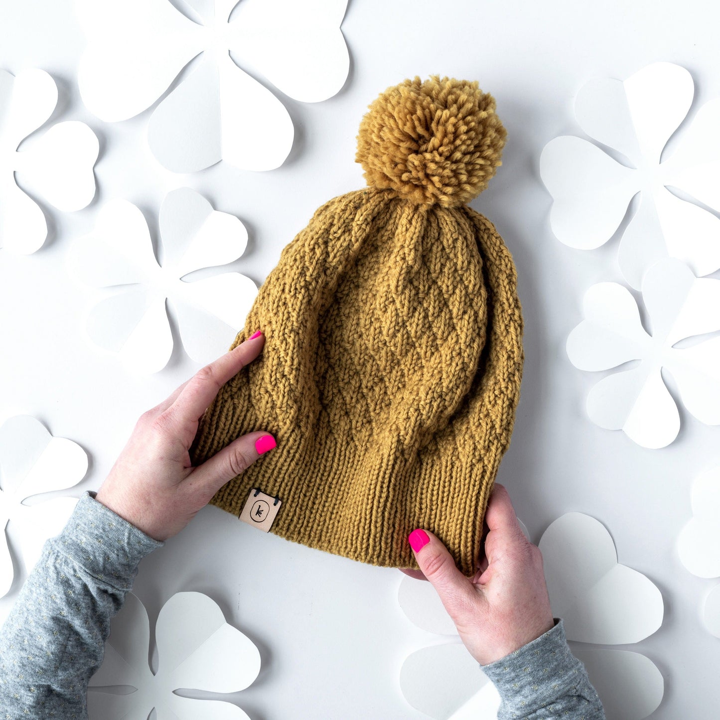 White hands with pink nail polish holding the March Hat pattern by Meghan Babin, knit out of Germantown in 708 honey with a large pom pom on a paper background with four leaf clover cut out shapes