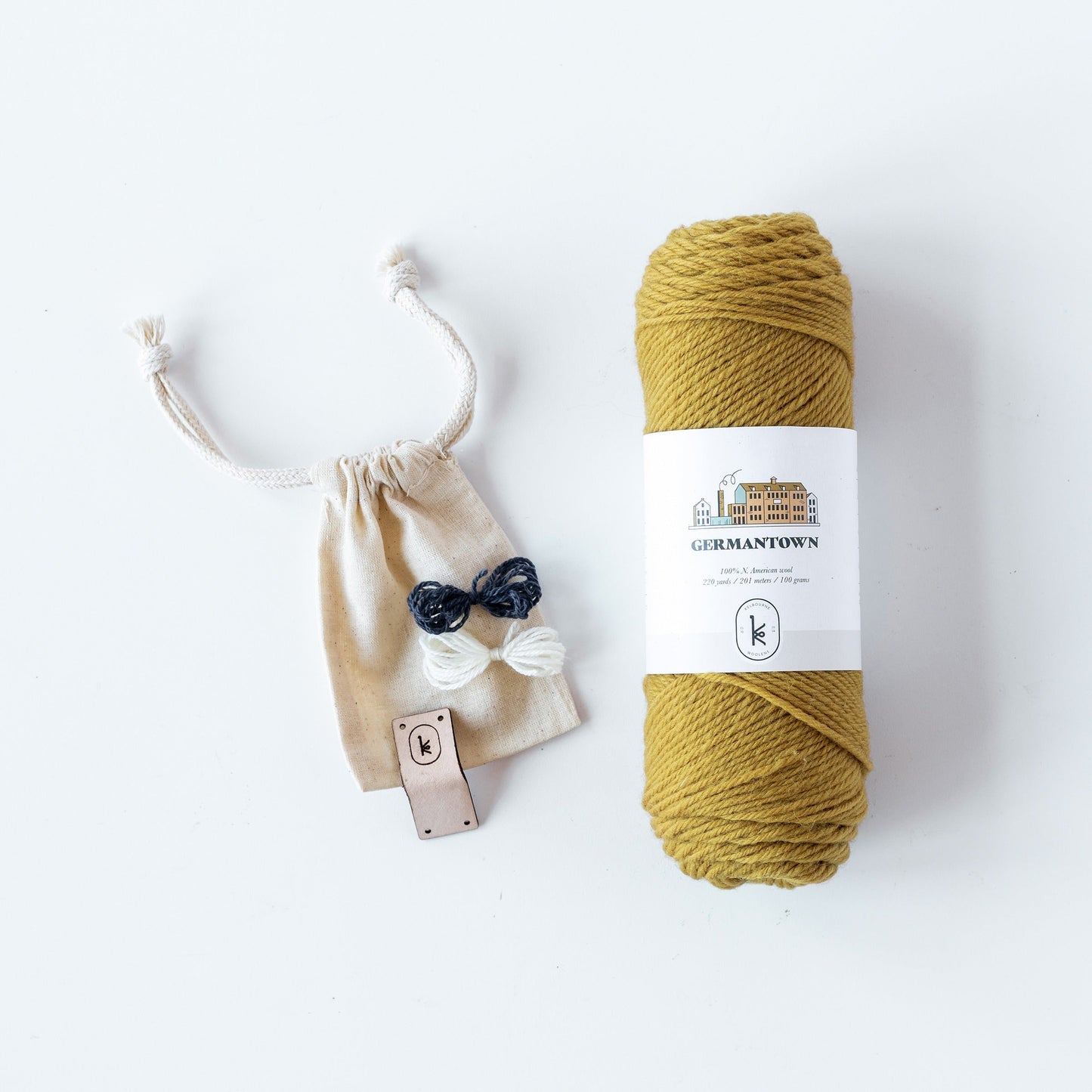 Flat lay of Kelbourne Woolens Germantown yarn skein in 708 honey with 2 small colorful yarn butterflies and one leather tag on a small muslin bag.
