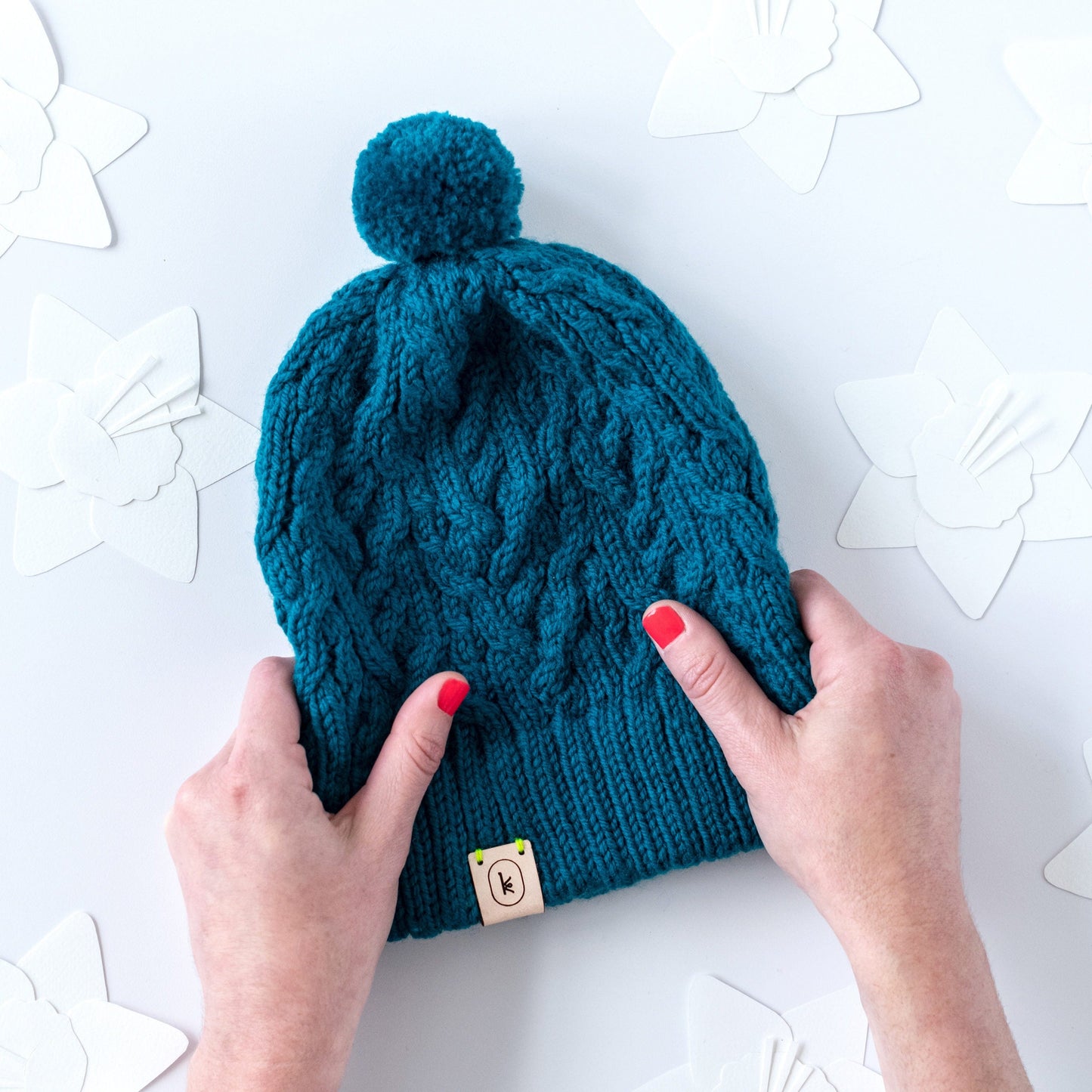 White hands with pink nail polish holding the intricately cabled May Hat pattern by Kate Gagnon Osborn in 440 peacock on a paper background with paper daffodil cut out shapes