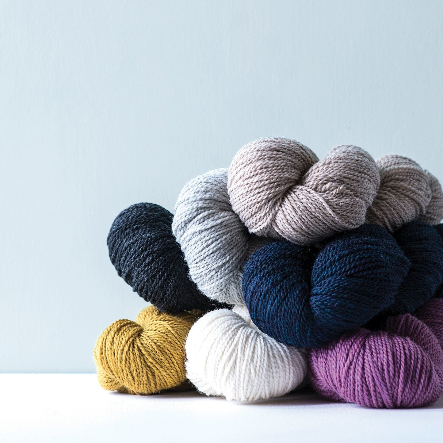 Stack of Kelbourne Woolens Scout heather wool yarn in natural gray, pink, gold on a white ground and light blue background