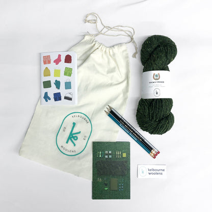 Overhead image of the contents of the March Year of Gifts kit: one skein of Kelbourne Woolens Lucky in pine, three pencils, a printed notebook, Kelbourne Woolens sticker, and project bag