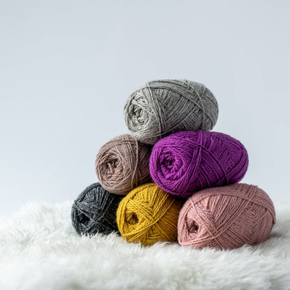 Stack of Navia Duo wool yarn in pink, gold, and natural gray on a shearling blanket and light gray background