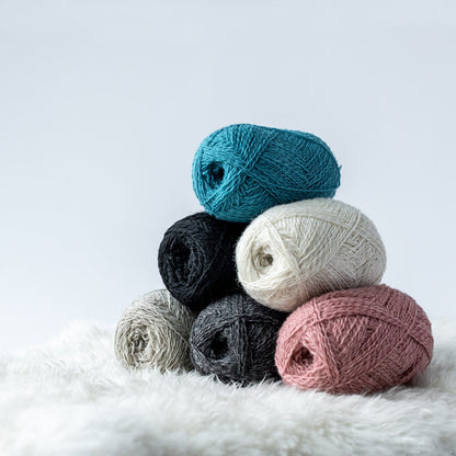 Stack of Navia Uno wool yarn in natural gray, pink, and turquoise on a shearling blanket and light gray background